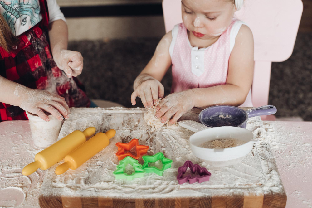 Two young children are playing with flour scattered on a wooden table with different shaped cutters and rolling pins